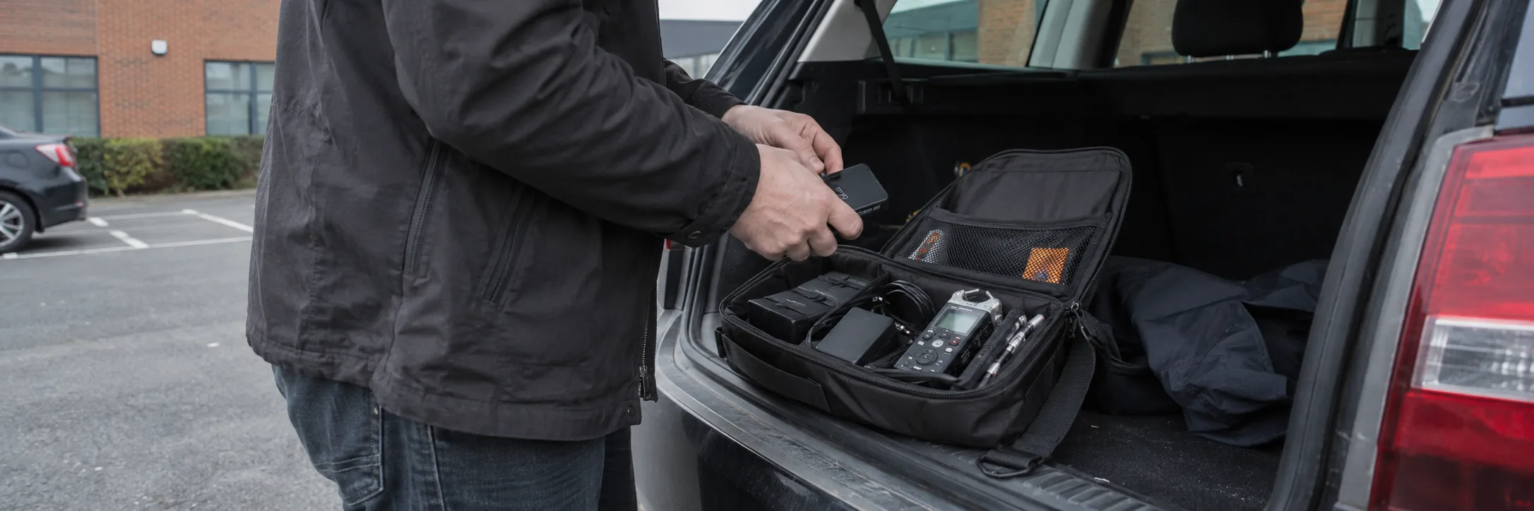 An investigator checking a compact technical field kit at the open boot of a vehicle.